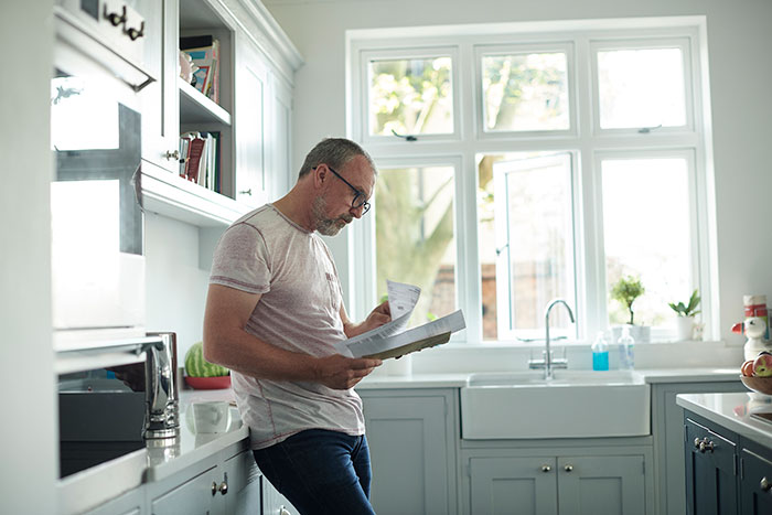 Man stood in a kitchen looking at a bill that has been received through the post Man stood in a kitchen looking at a bill that has been received through the post
